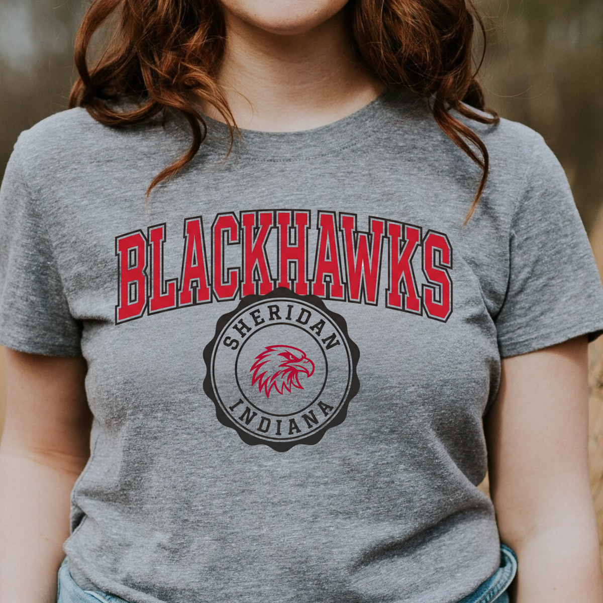 Close-up of a woman wearing a grey t-shirt featuring the classic collegiate Sheridan Blackhawks wordmark and team seal.