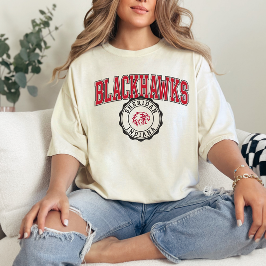 Woman in a cream-colored relaxed-fit t-shirt showcasing the Heritage Collegiate Sheridan Blackhawks graphic.