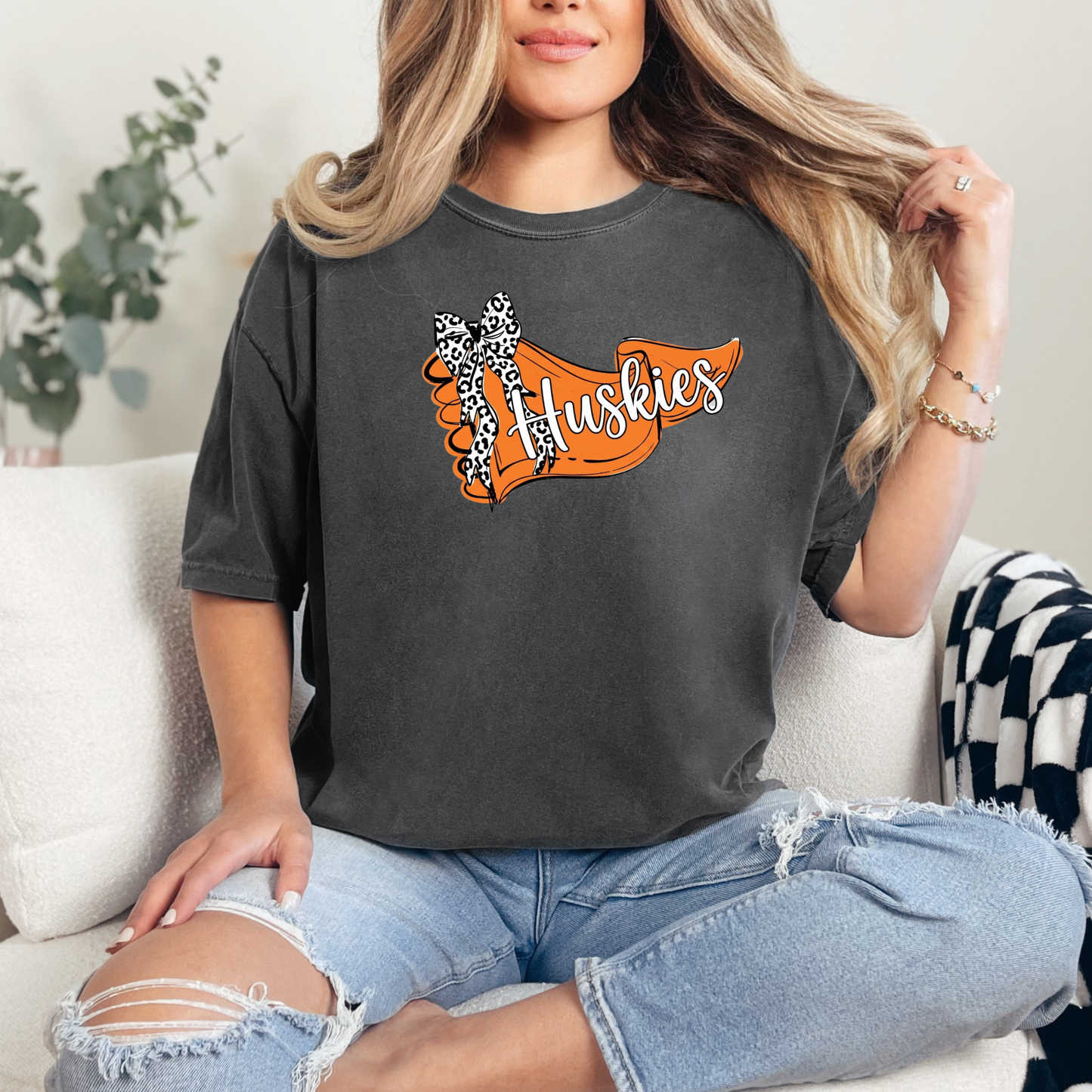A person wearing a dark grey t-shirt featuring an orange banner design with 'Huskies' text and a leopard-print bow.