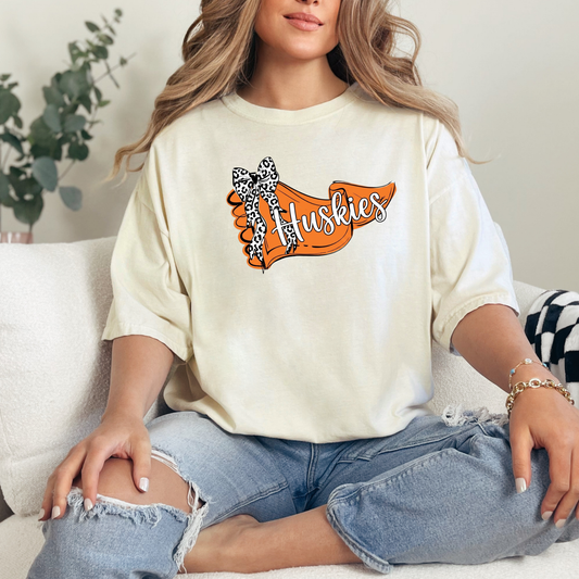 A person sitting on a couch wearing a cream-colored t-shirt featuring an orange banner design with 'Huskies' text and a leopard-print bow.