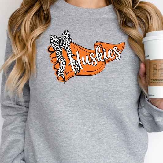 A person wearing a soft grey crewneck sweatshirt adorned with a custom orange banner featuring 'Huskies' text and a trendy leopard-print bow, holding a disposable coffee cup.