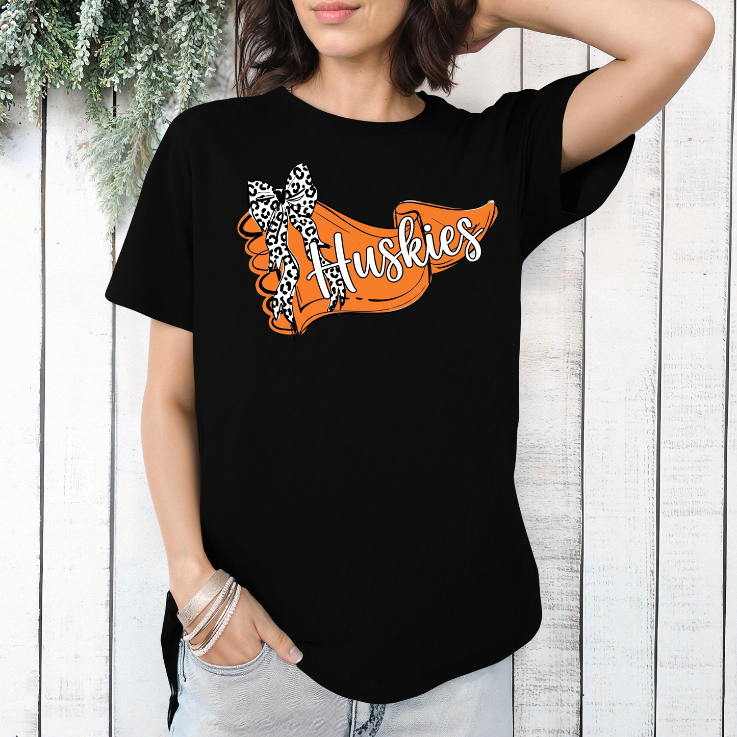 A person wearing a black t-shirt featuring an orange banner design with 'Huskies' text and a leopard-print bow, posed in front of a white wooden wall.