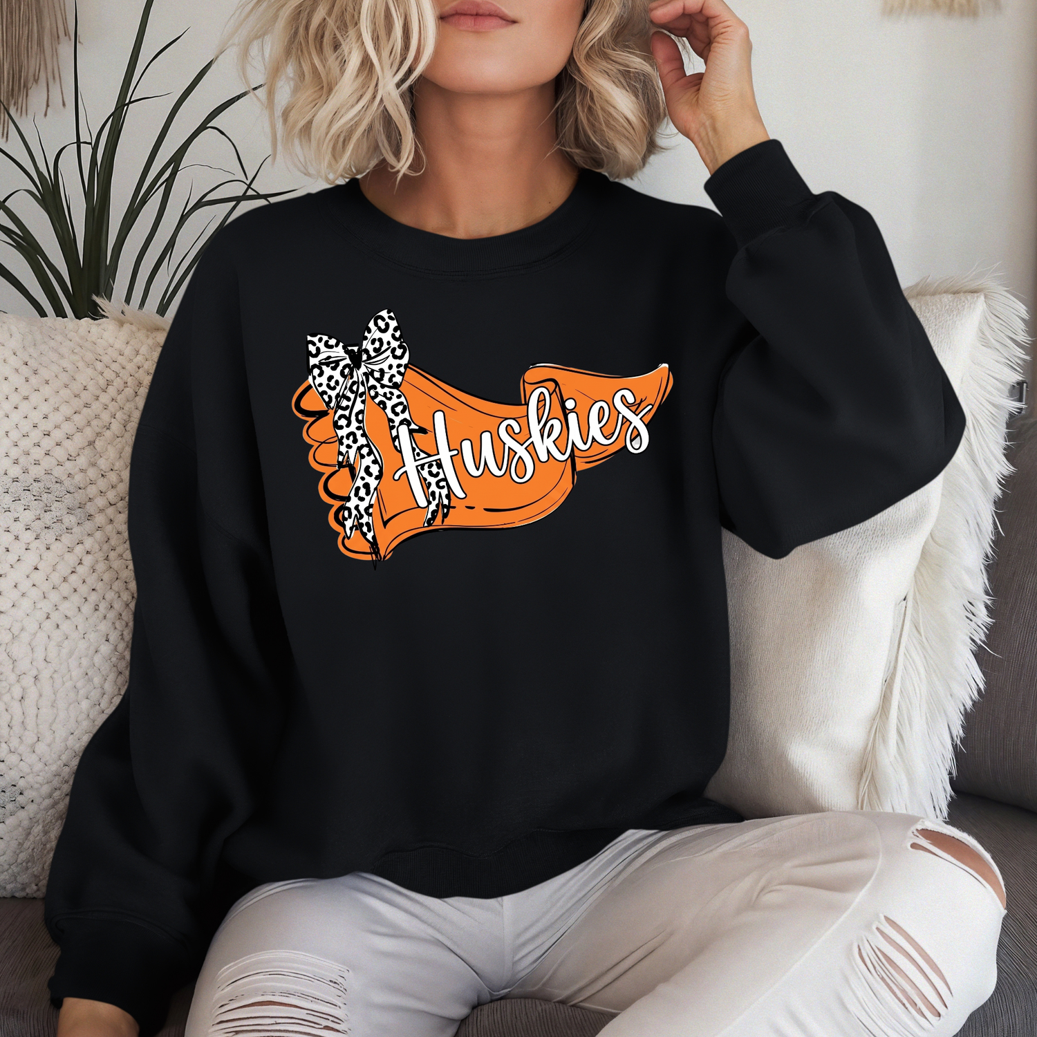 A person lounging on a couch wearing a black crewneck sweatshirt that displays an orange pennant-style banner graphic with 'Huskies' in white script and a leopard-print bow.
