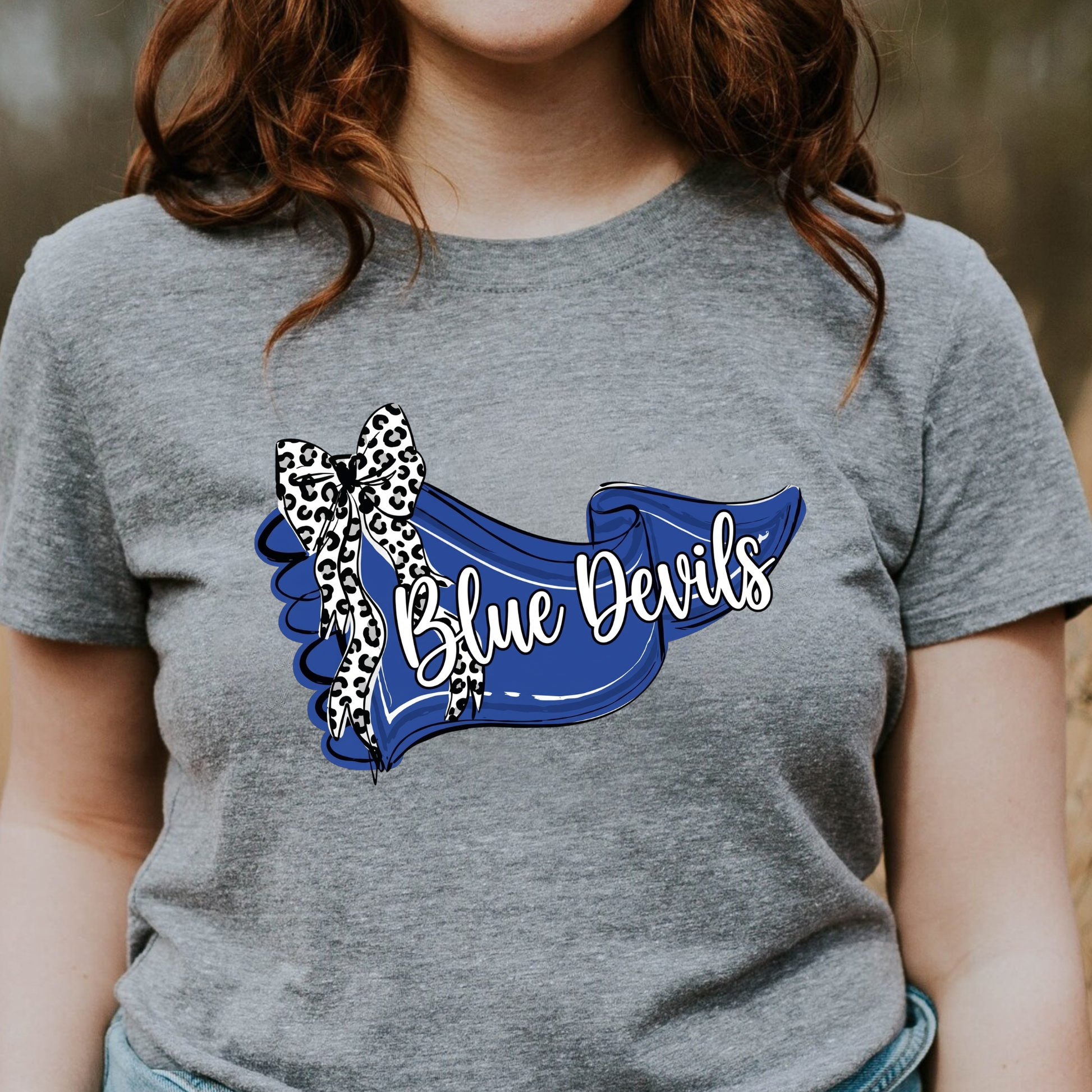 Woman wearing a grey graphic t-shirt featuring a blue 'Blue Devils' banner design with a leopard-print bow."