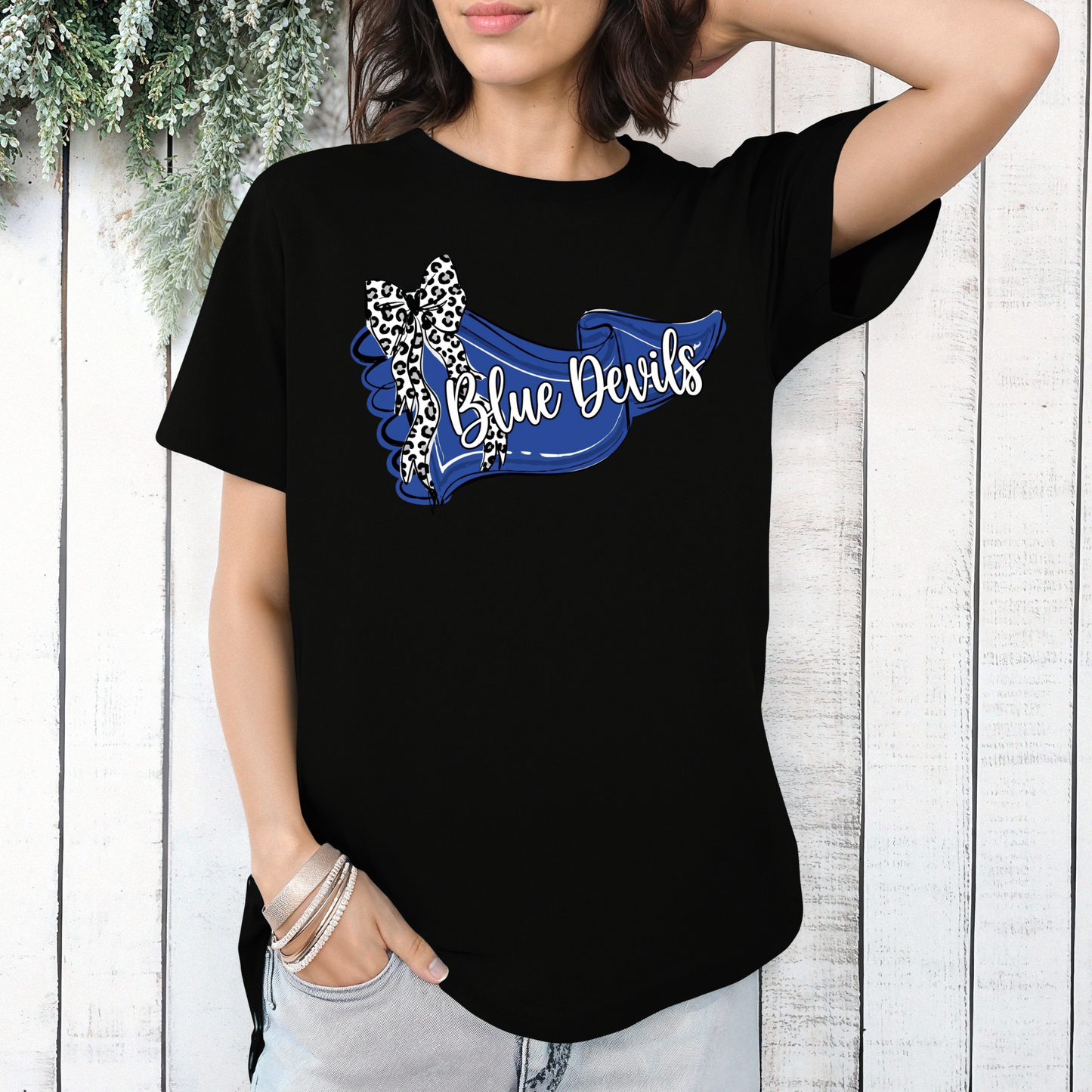 Woman wearing a black graphic t-shirt featuring a blue 'Blue Devils' banner design with a leopard-print bow, styled against a white wooden wall.