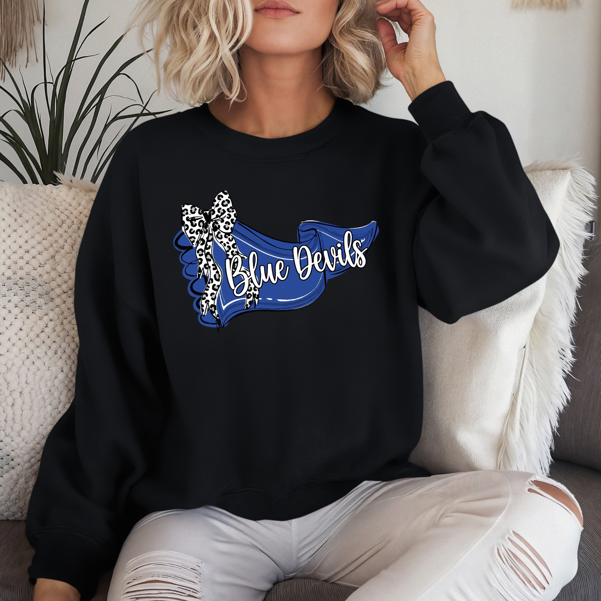 Woman sitting on a couch wearing a black crewneck sweatshirt featuring a blue 'Blue Devils' banner design with a leopard-print bow.