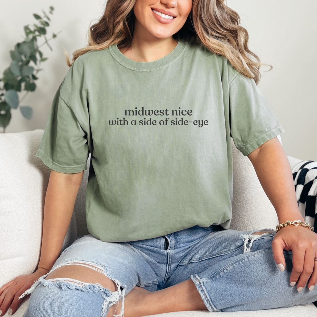 Woman wearing a Bay graphic tee that says ‘midwest nice with a side of side-eye,’ styled with ripped jeans in a cozy living room.