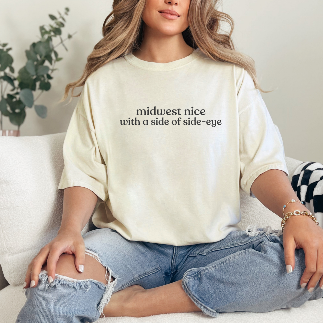 Woman wearing an ivory graphic tee that says ‘midwest nice with a side of side-eye,’ styled with ripped jeans in a cozy living room.