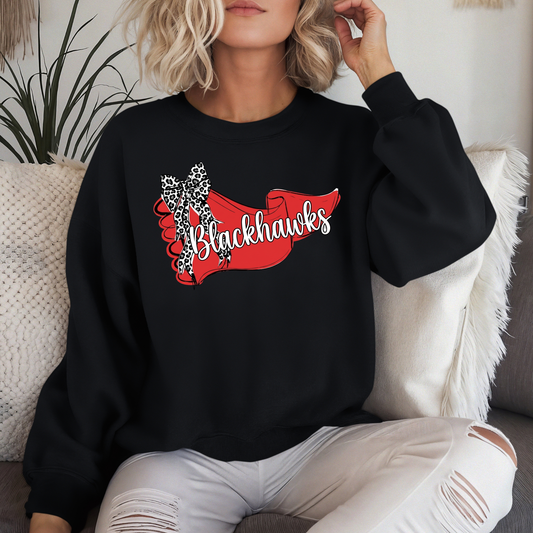 Woman wearing a black crewneck sweatshirt featuring a Sheridan Blackhawks banner and leopard-print bow graphic, sitting on a couch.