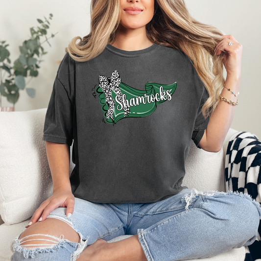 A person wearing a dark grey t-shirt featuring a green banner design with 'Shamrocks' text and a leopard-print bow.