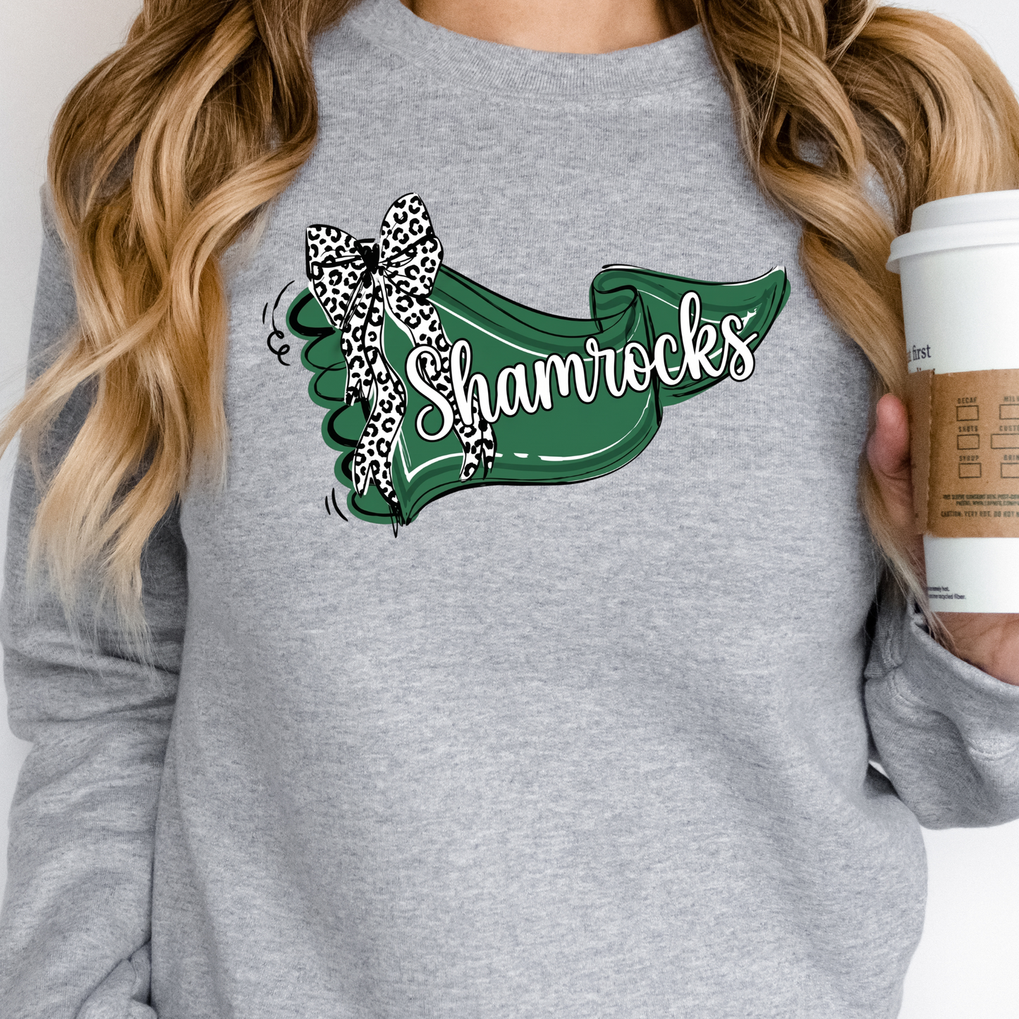 A person wearing a soft grey crewneck sweatshirt featuring a green banner graphic with 'Shamrocks' text and a leopard-print bow, holding a coffee cup.