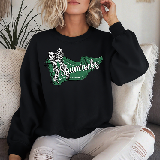 A person lounging on a couch wearing a black crewneck sweatshirt that displays a green banner graphic with 'Shamrocks' in white script and a leopard-print bow.