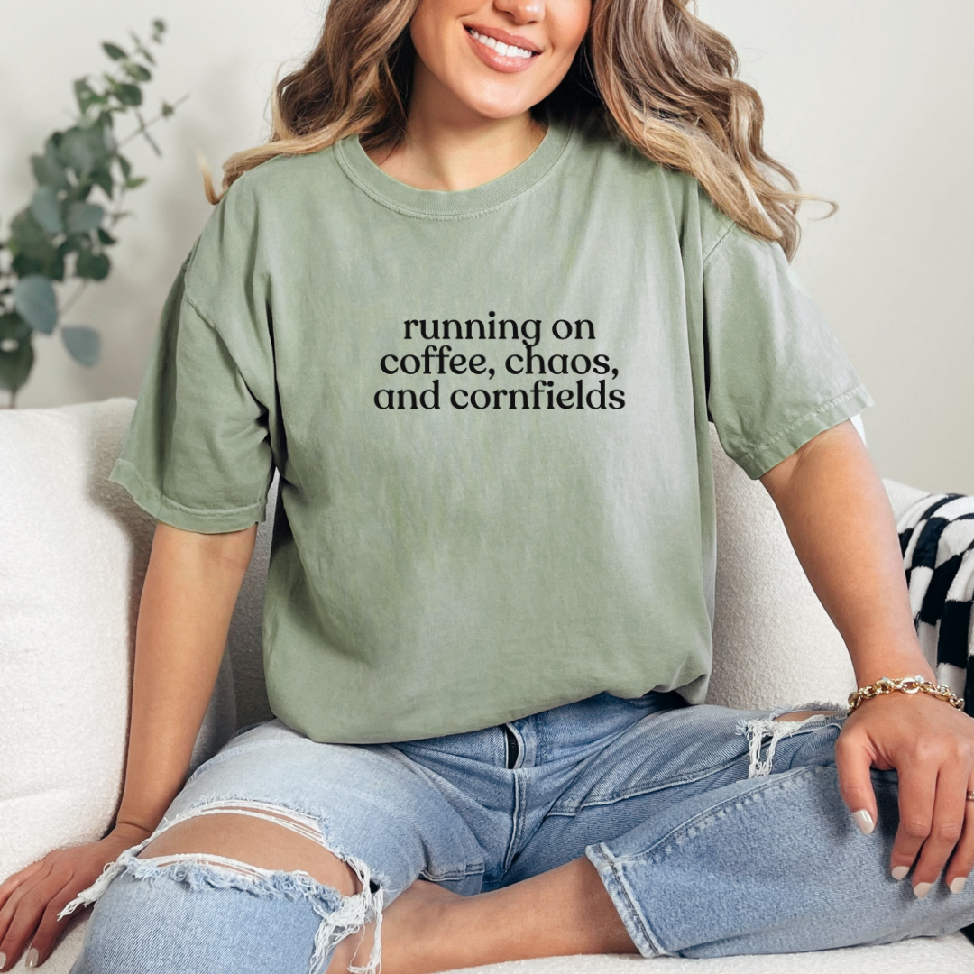 Woman sitting on a couch wearing a Bay Comfort Colors t-shirt that says ‘running on coffee, chaos, and cornfields’ in black text