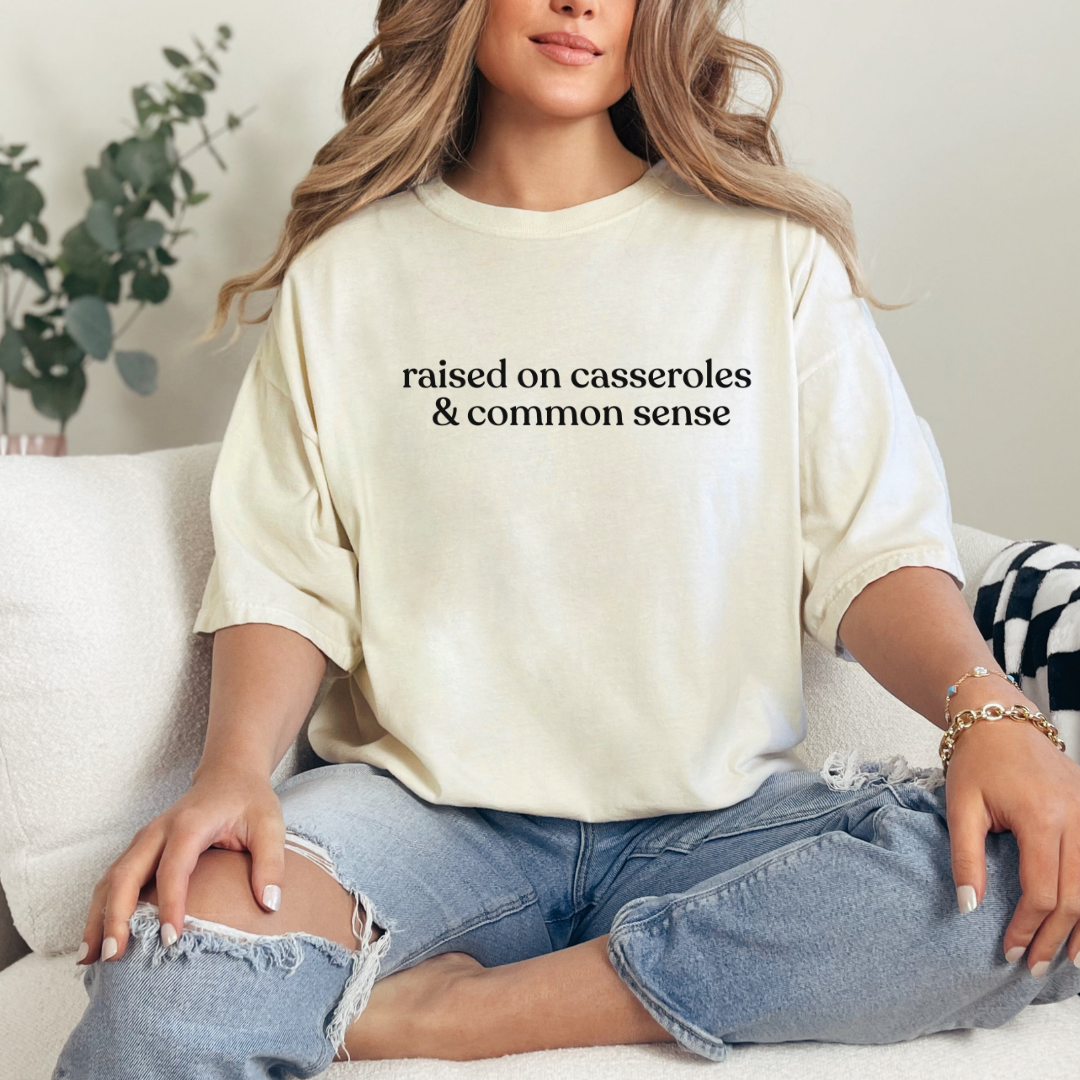 A woman sitting casually on a sofa wearing an ivory Comfort Colors T-shirt with the phrase ‘raised on casseroles & common sense’ printed in black. Cozy, relaxed home setting.