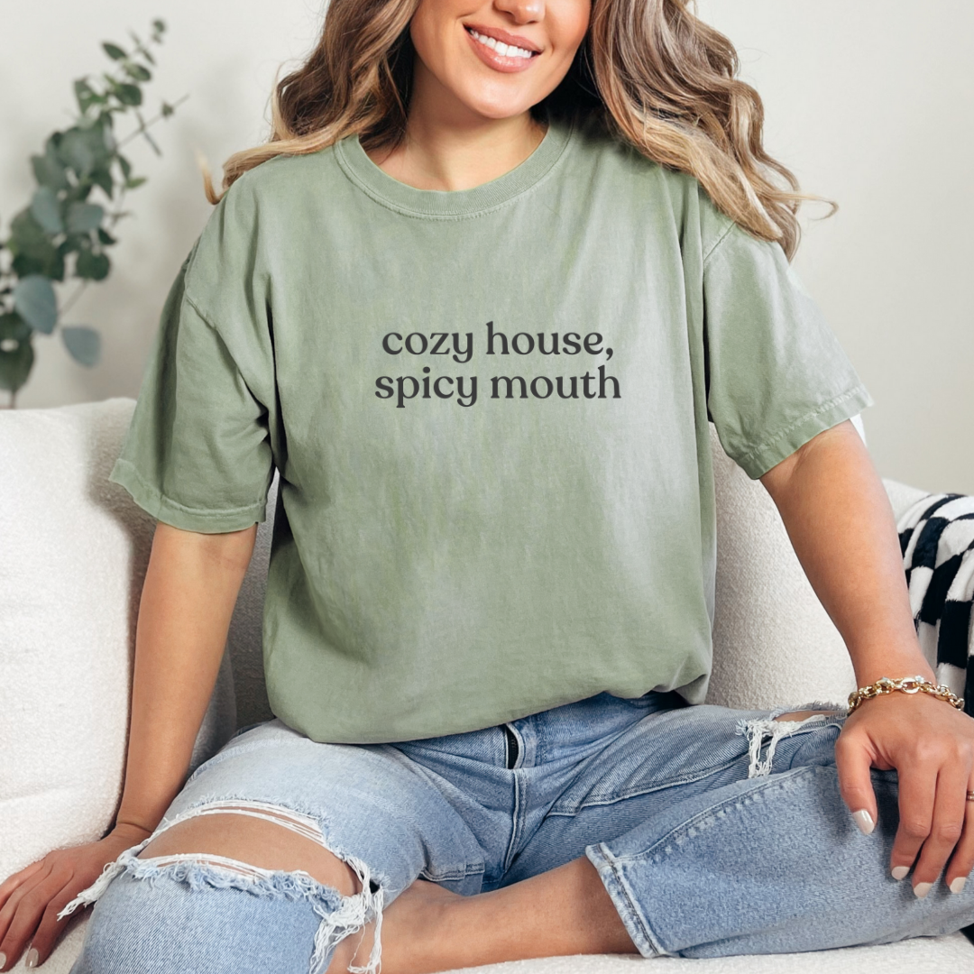 Woman wearing a Bay graphic tee with the phrase “cozy house, spicy mouth,” seated casually on a sofa in a relaxed home setting.