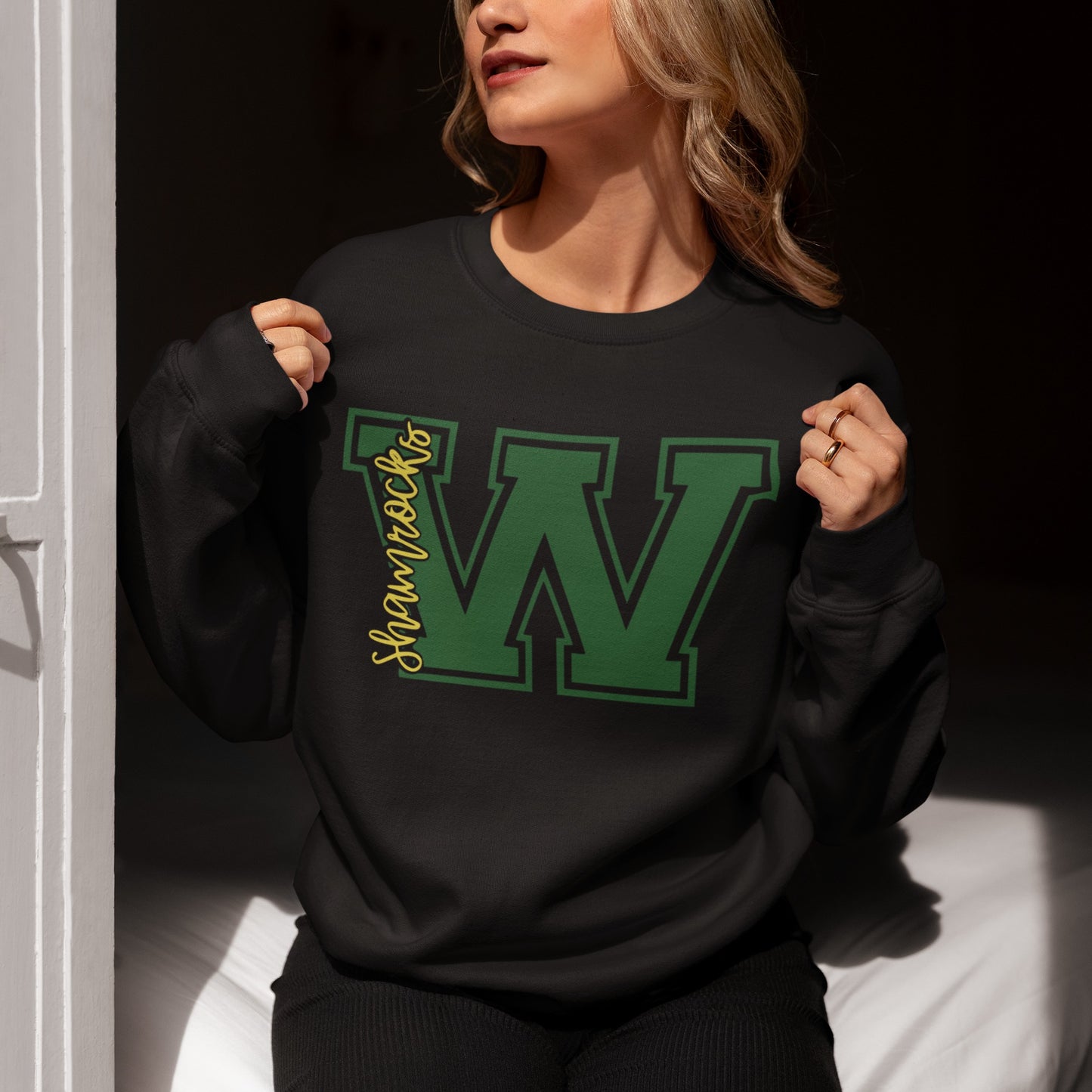 Woman sitting in soft natural light wearing a black Westfield Shamrocks varsity ‘W’ crewneck sweatshirt with green and yellow lettering.
