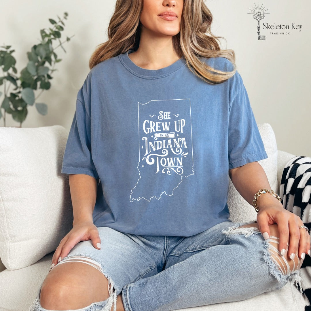 Woman wearing a Blue Jean Comfort Colors tee with a white “She Grew Up in an Indiana Town” Indiana state outline graphic.