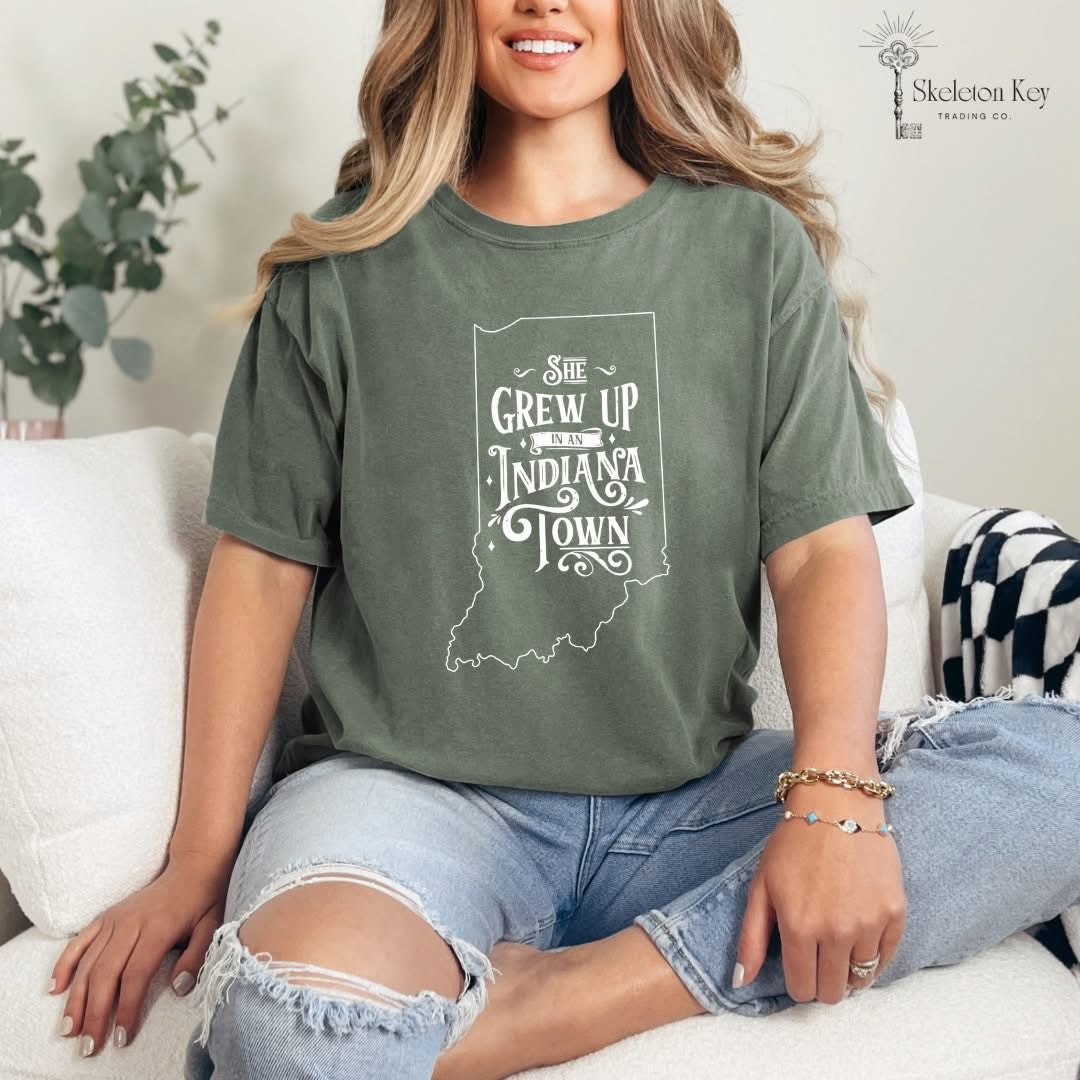 Woman wearing a Sage Comfort Colors tee with a white “She Grew Up in an Indiana Town” Indiana state outline graphic.
