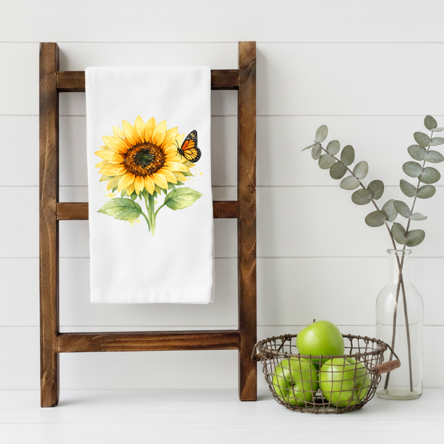 Watercolor sunflower tea towel with monarch butterfly hanging on a wooden ladder in a farmhouse-style kitchen setting.