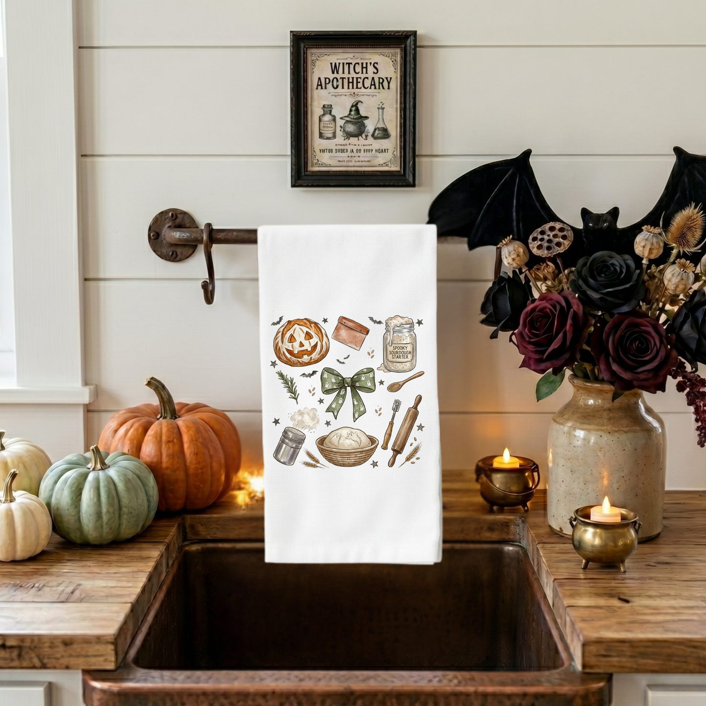 Decorative kitchen towel with Halloween-themed design hanging over a sink, surrounded by pumpkins and candles.