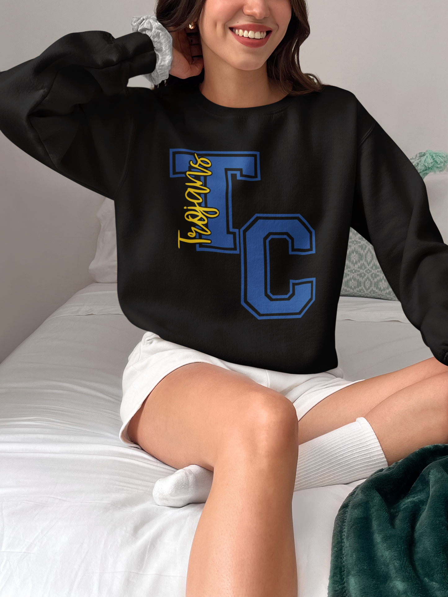 Woman wearing black Tri-Central Trojans varsity TC crewneck sweatshirt while sitting on a bed