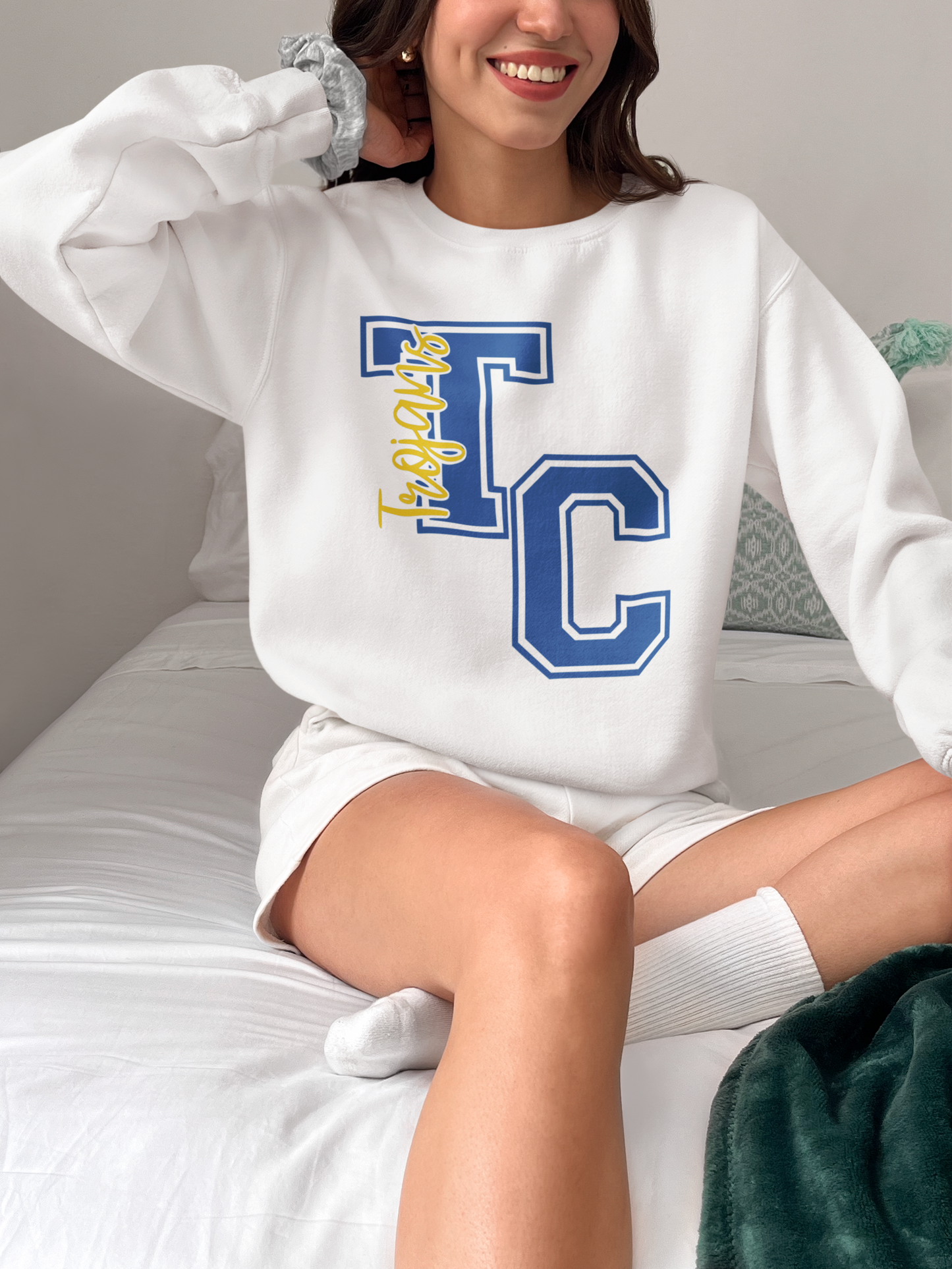 Woman wearing white Tri-Central Trojans varsity TC crewneck sweatshirt while sitting on a bed