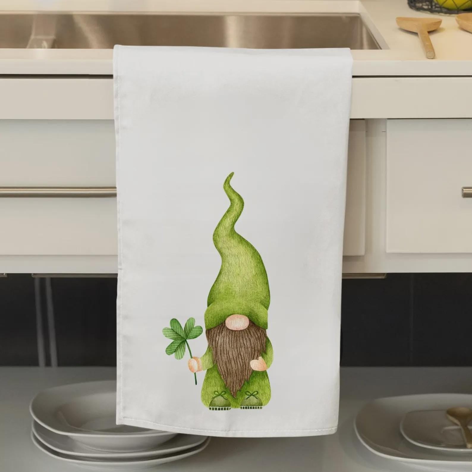 May include: White kitchen towel featuring a watercolor illustration of a green gnome holding a four-leaf clover. The gnome has a long, curly green hat, a brown beard, and green clothing. The towel is hanging on a cabinet door.