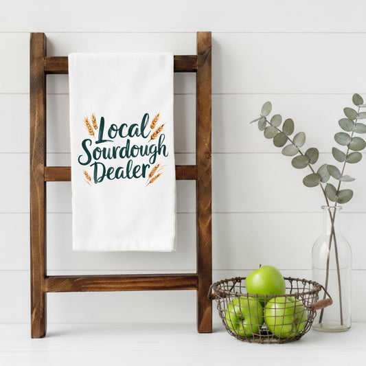 White towel with 'Local Sourdough Dealer' text on a wooden rack, with apples and a vase in the background.