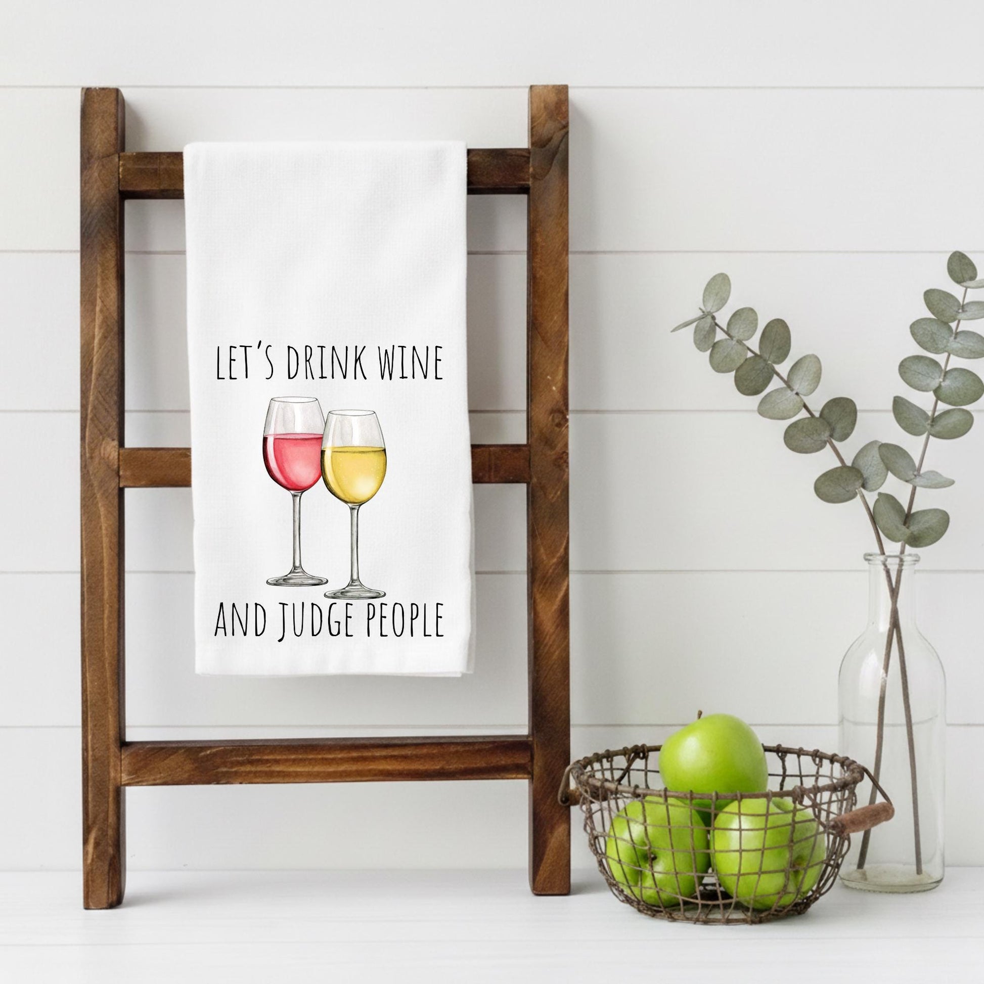 Tea towel with 'Let's drink wine and judge people' text on a wooden ladder, with apples and a vase in the background.