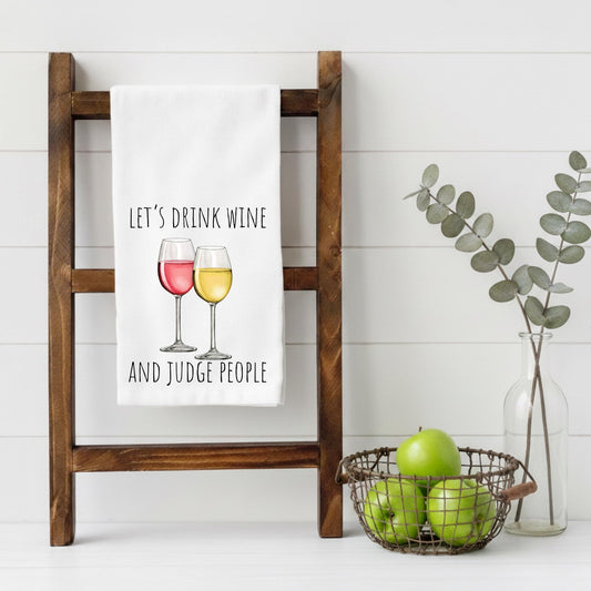 Tea towel with 'Let's drink wine and judge people' text on a wooden ladder, with apples and a vase in the background.