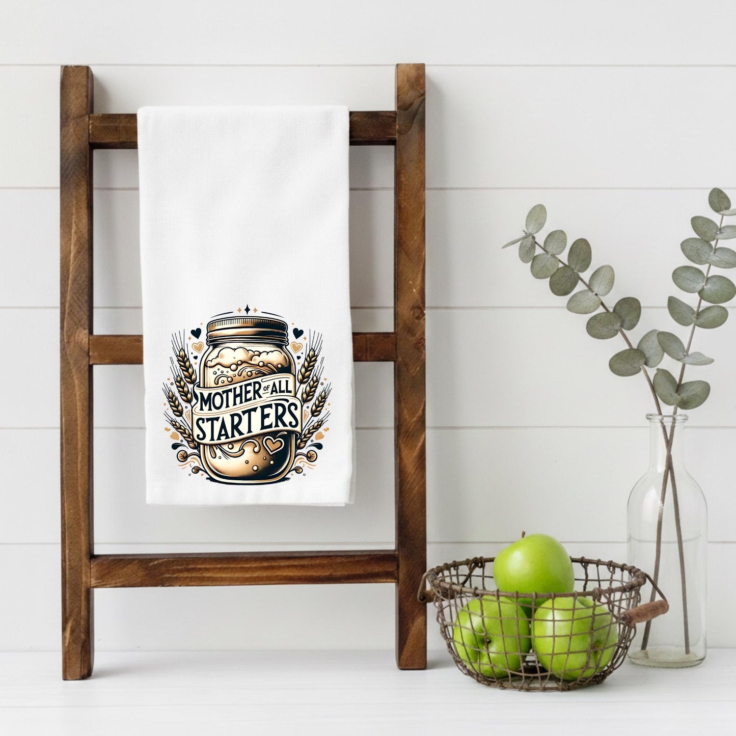 White towel with 'Mother of All Starters' design on a wooden ladder, with a basket of apples and a vase with greenery in the background.
