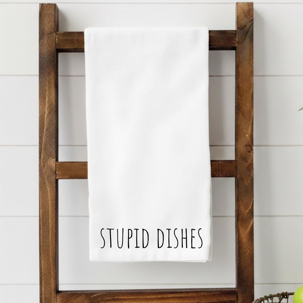 White towel with 'STUPID DISHES' text on a wooden towel rack against a white wall.