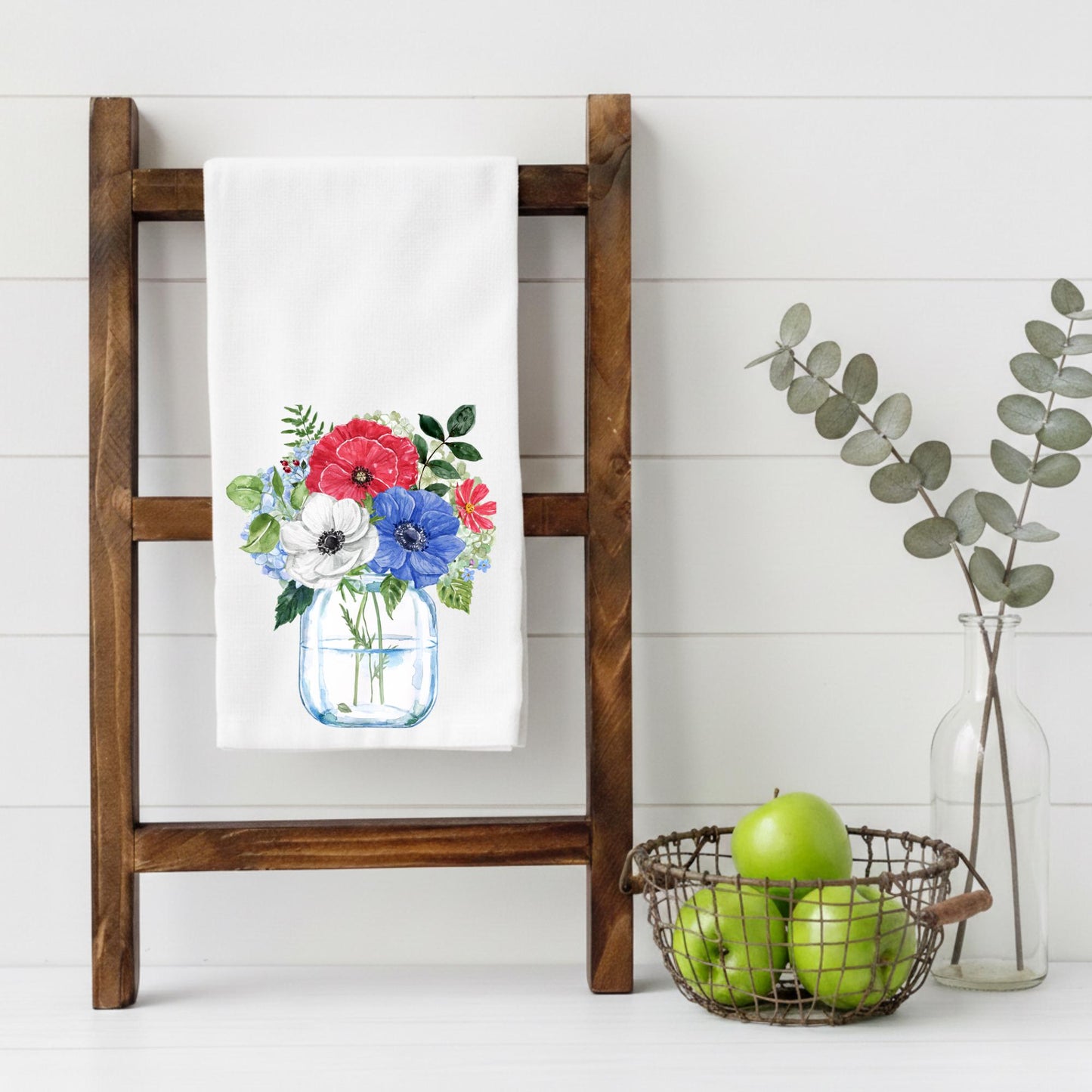 Floral towel on a wooden ladder with a basket of apples and a vase of eucalyptus.