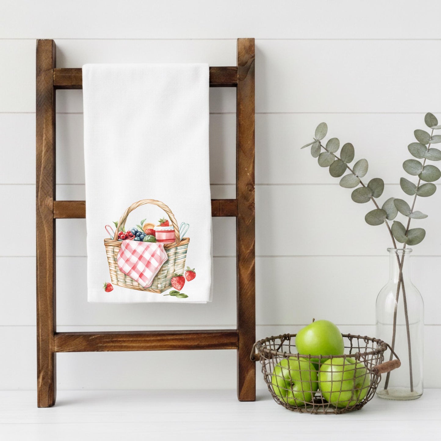 Wooden ladder with a towel featuring a picnic basket design, green apples in a wire basket, and a vase with eucalyptus leaves on a white background.