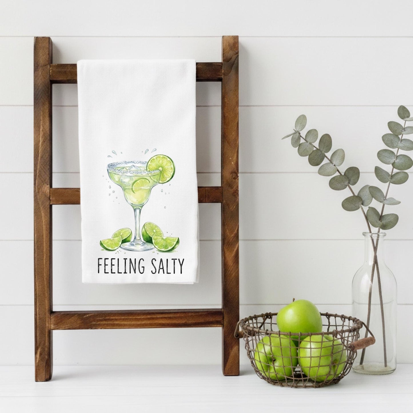 Kitchen towel with a margarita design hanging on a wooden rack, surrounded by apples and eucalyptus.