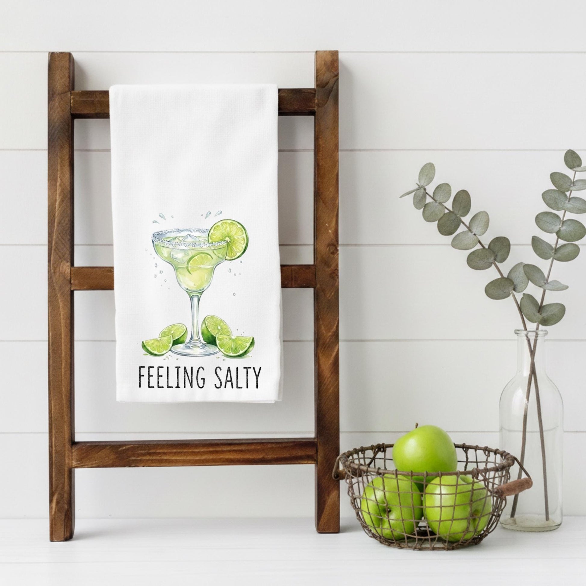 Kitchen towel with a margarita design hanging on a wooden rack, surrounded by apples and eucalyptus.