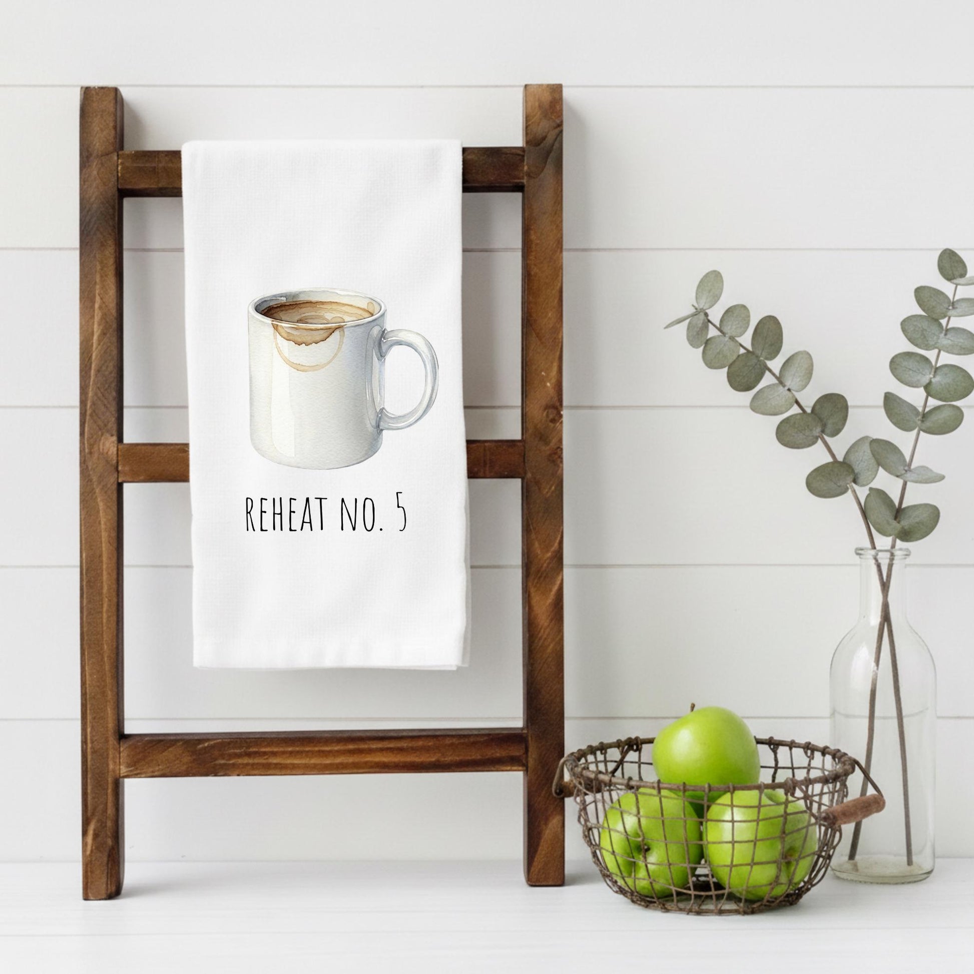 Wooden towel rack with a towel featuring a coffee cup design, basket of apples, and vase with eucalyptus on a white background.