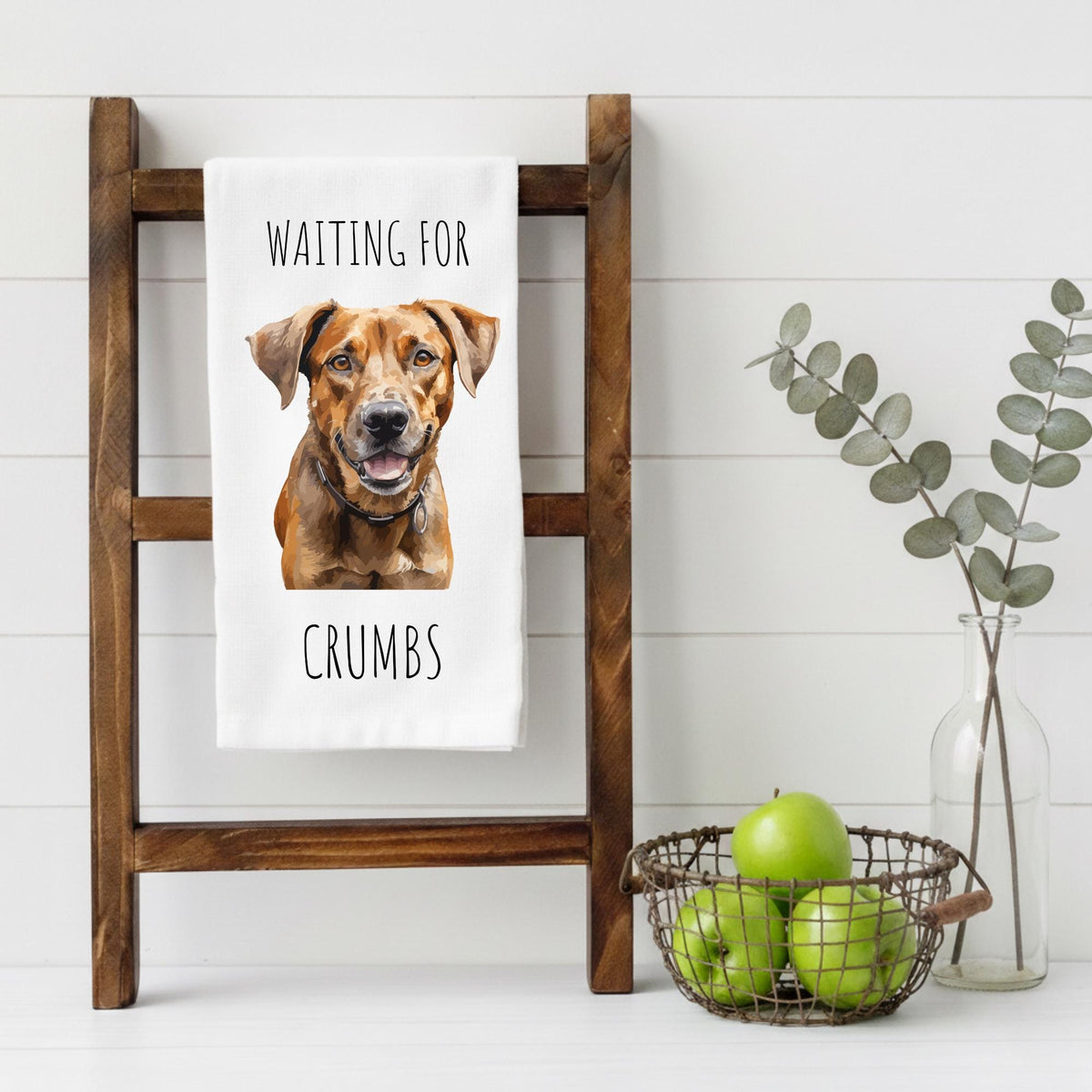 Tea towel with a dog illustration and text on a wooden ladder, with a basket of apples and a vase in the background.