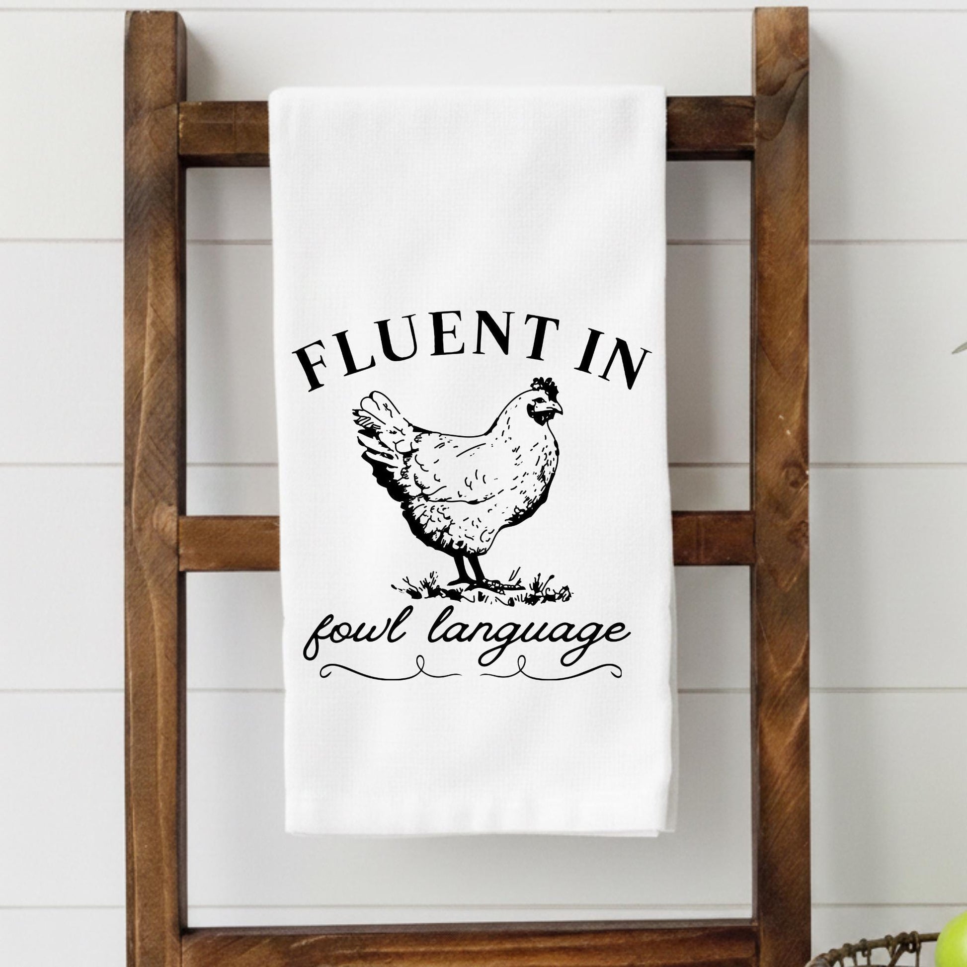 White towel with black text and chicken illustration on a wooden ladder against a white wall.