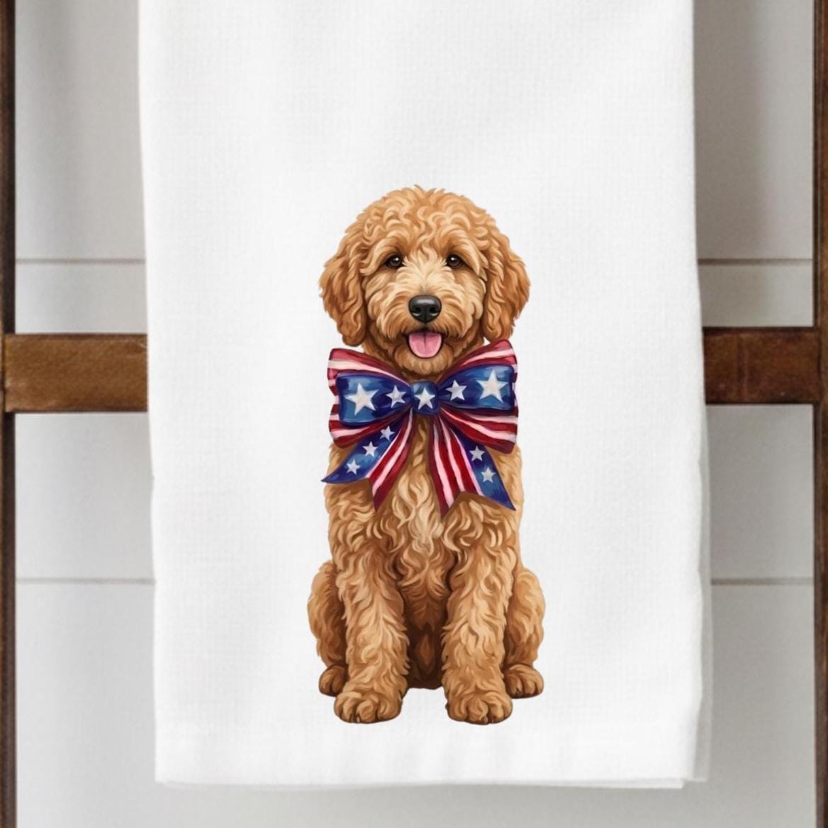 A white towel hanging on a wooden rack, featuring a printed illustration of a golden doodoodle wearing a patriotic red, white, and blue star-patterned bow tie.