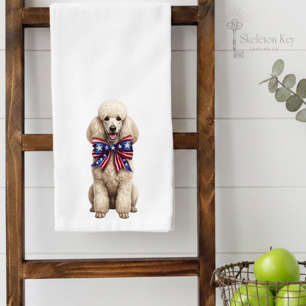 A white kitchen towel hanging on a wooden ladder, printed with an illustration of a poodle wearing a red, white, and blue patriotic bow. The background is a white shiplap wall with a decorative eucalyptus branch and a wire basket of green apples.
