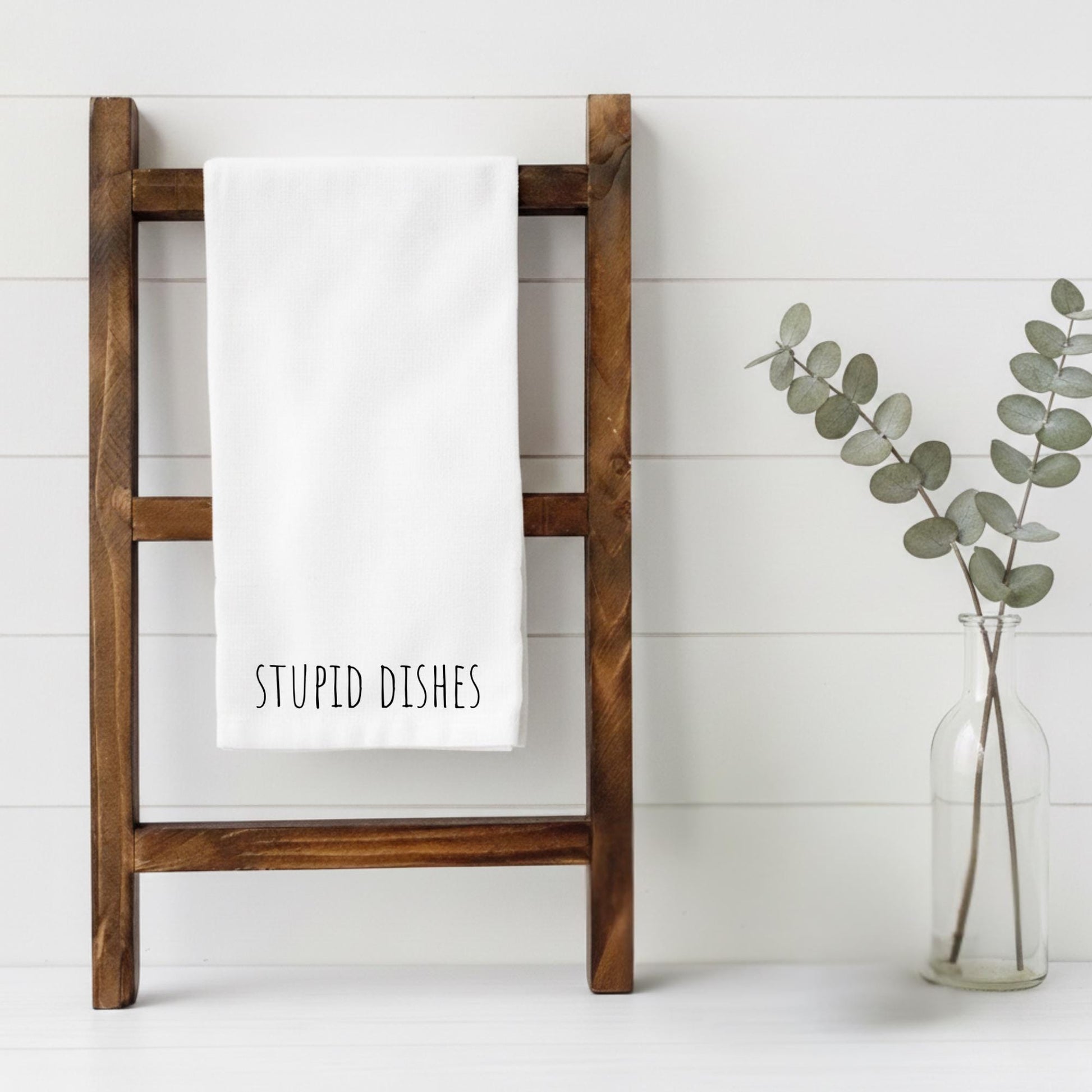 Wooden towel rack with a white towel labeled 'STUPID DISHES' next to a vase with eucalyptus on a white background