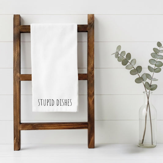 Wooden towel rack with a white towel labeled 'STUPID DISHES' next to a vase with eucalyptus on a white background