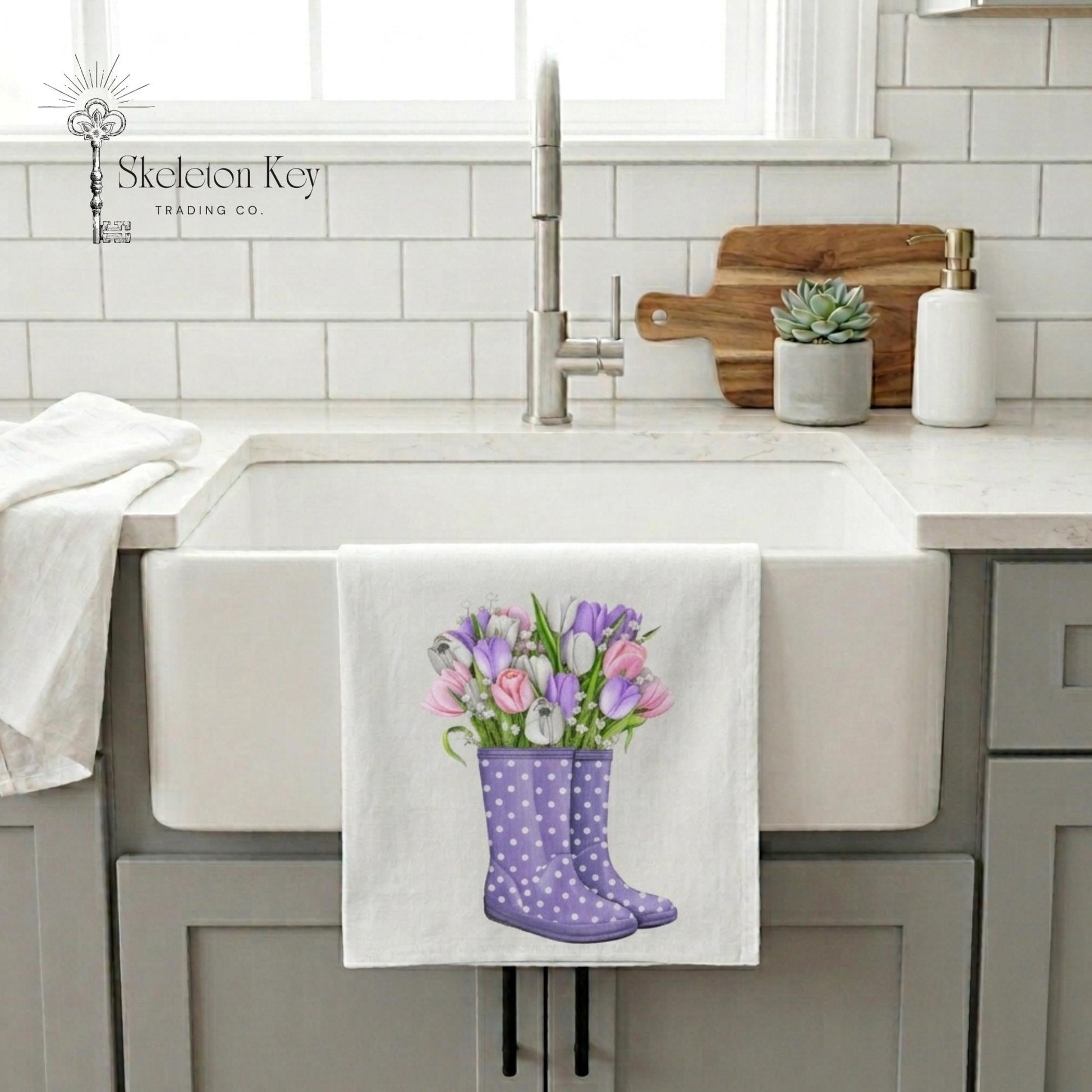 Kitchen sink with a towel hanging on it featuring purple rain boots and flowers, brand 'Skeleton Key Trading Co.' visible.