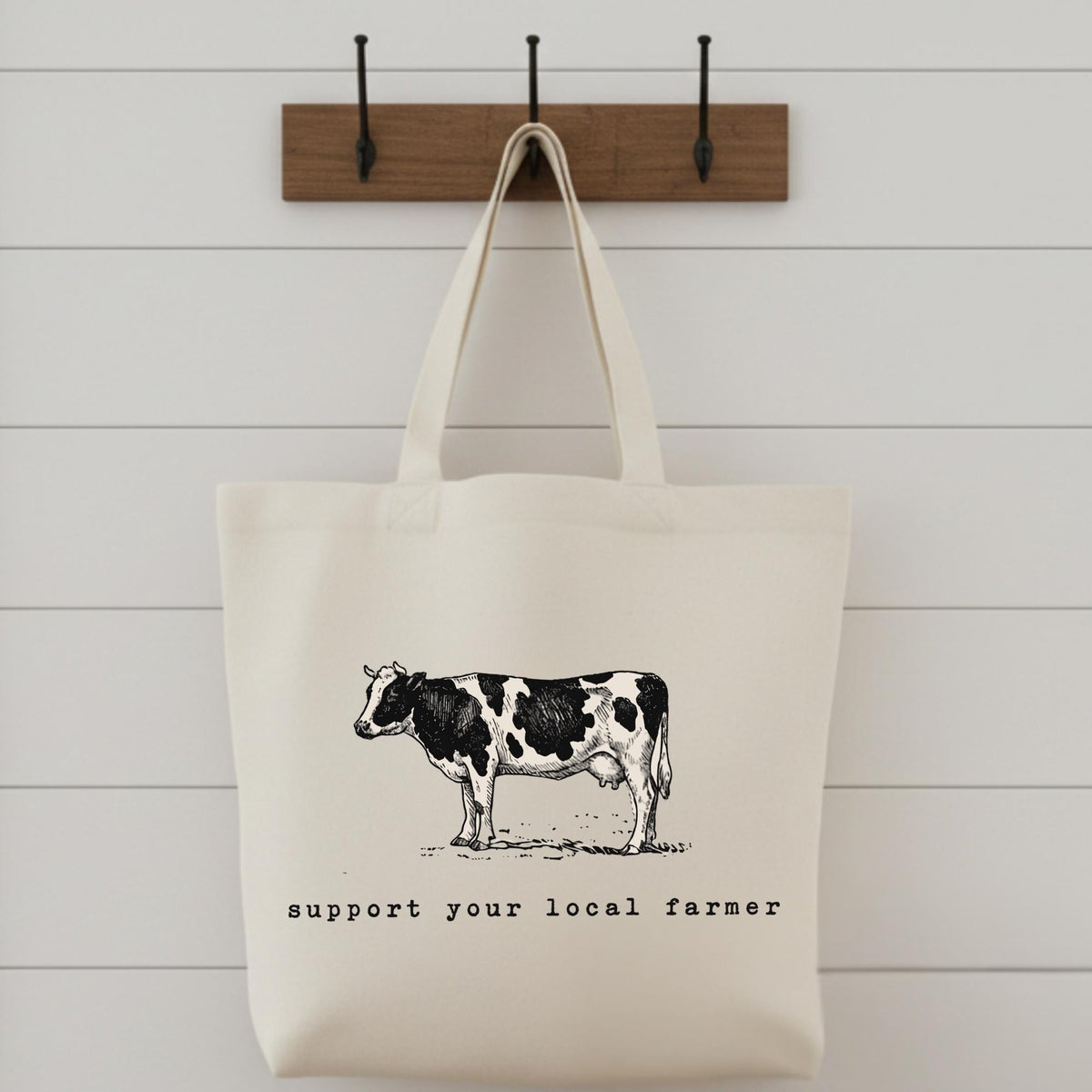 An off-white canvas tote bag with a black-and-white cow illustration and the text 'support your local farmer' hanging from a wooden wall hook against a white shiplap wall.