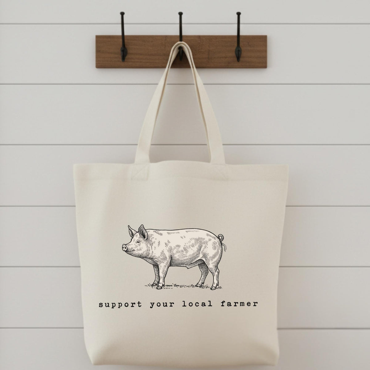 Beige tote bag with pig illustration and 'support your local farmer' text on a wooden hook against a white wall.