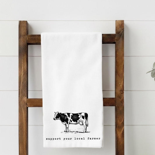 White towel with a cow illustration and text on a wooden ladder against a white wall.
