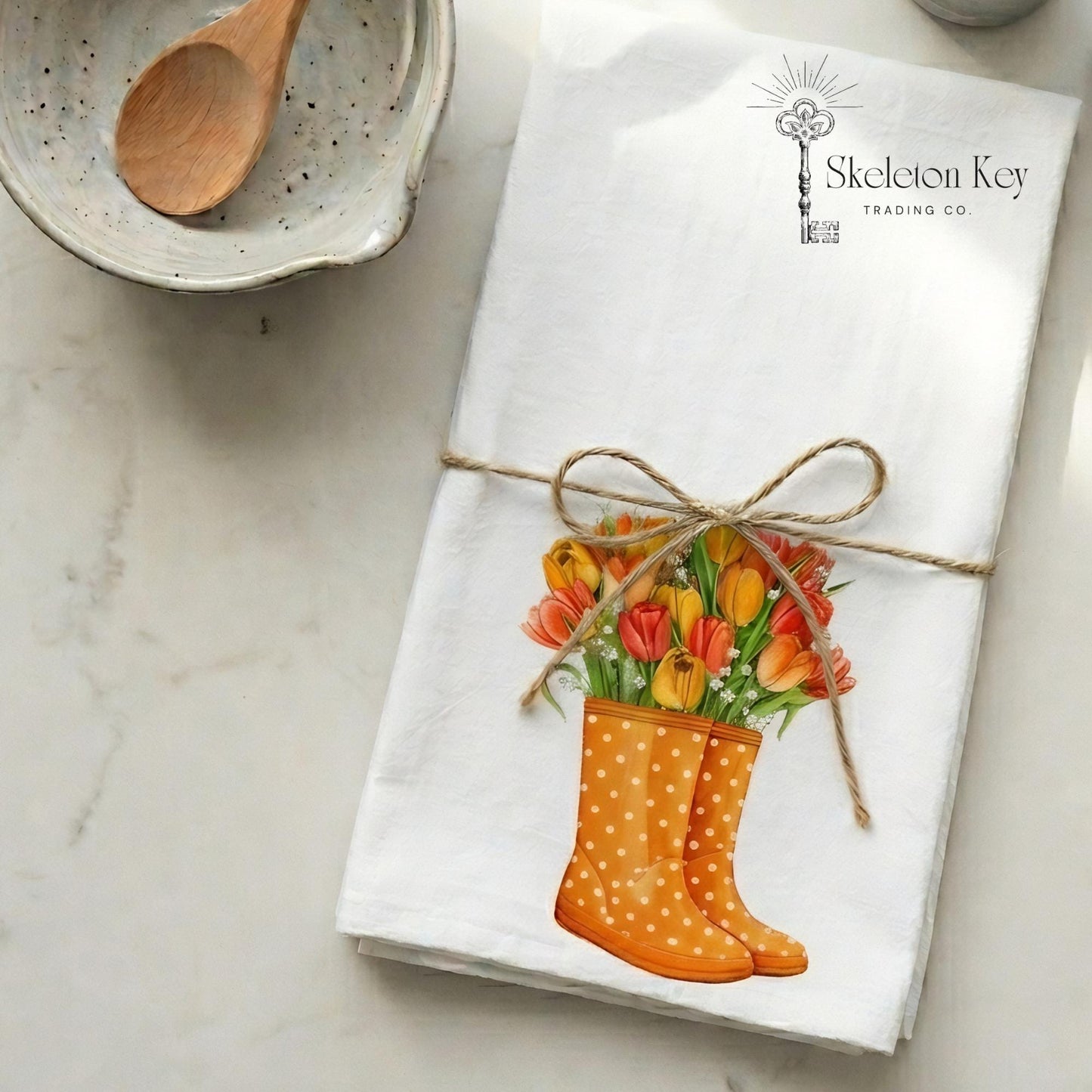 Gift display of a flour sack tea towel with an illustration of orange rainboots with colorful flowers.