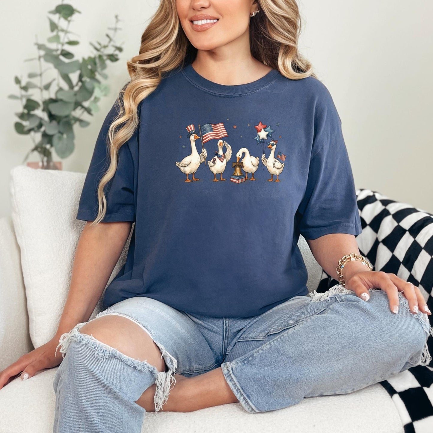 Woman wearing a navy blue t-shirt with a graphic design of ducks and American flags, sitting on a couch.