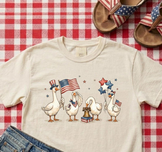 Beige t-shirt with duck illustrations holding American flags and stars on a red and white checkered tablecloth.
